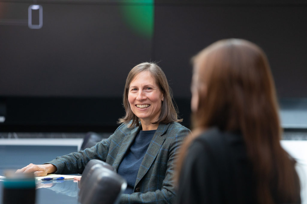 011822_Viking-CT_Candid-Office_057_Lo-Res A woman sits at a table and smiles at another colleague sitting next to her.