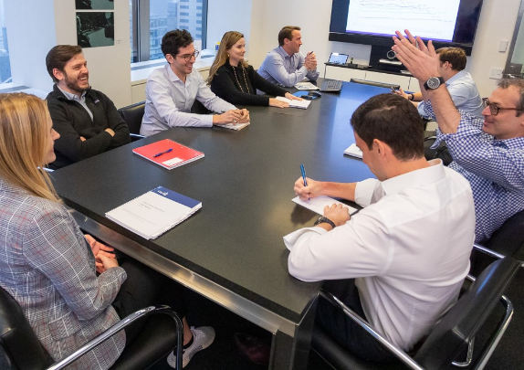 Colleagues gather around a meeting room table as one man gestures with his hands.