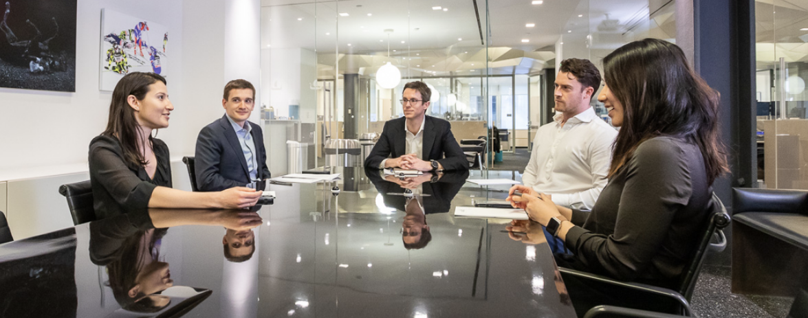 Mask Group 35 Five colleagues sit around a long table in a conference room with glass walls.
