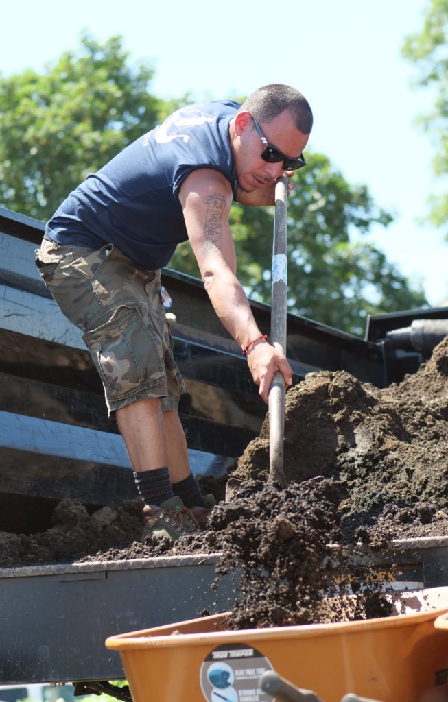 Mike gardening_cropped A man stands in the back of a pick up truck and shovels dirt into a wheelbarrow.