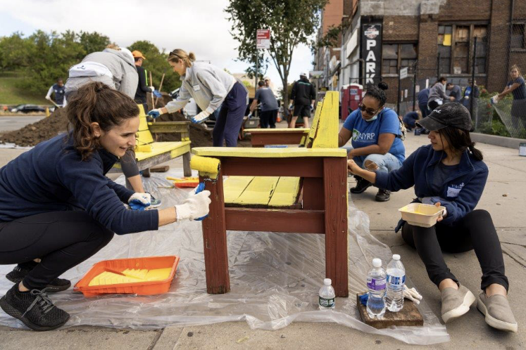 Three colleagues paint a park bench with yellow paint.