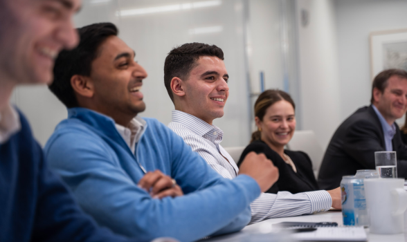 Colleagues sit next to each other at a conference table and smile to someone off camera.